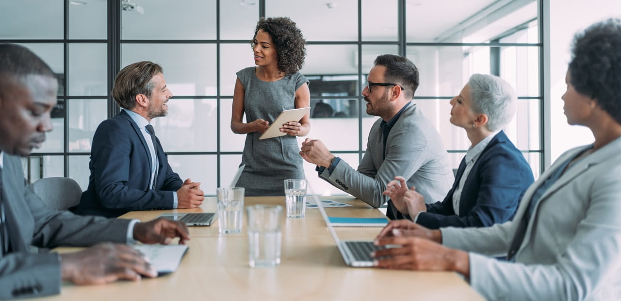 Female leader standing and speaking to a group of professionals sitting at a conference table