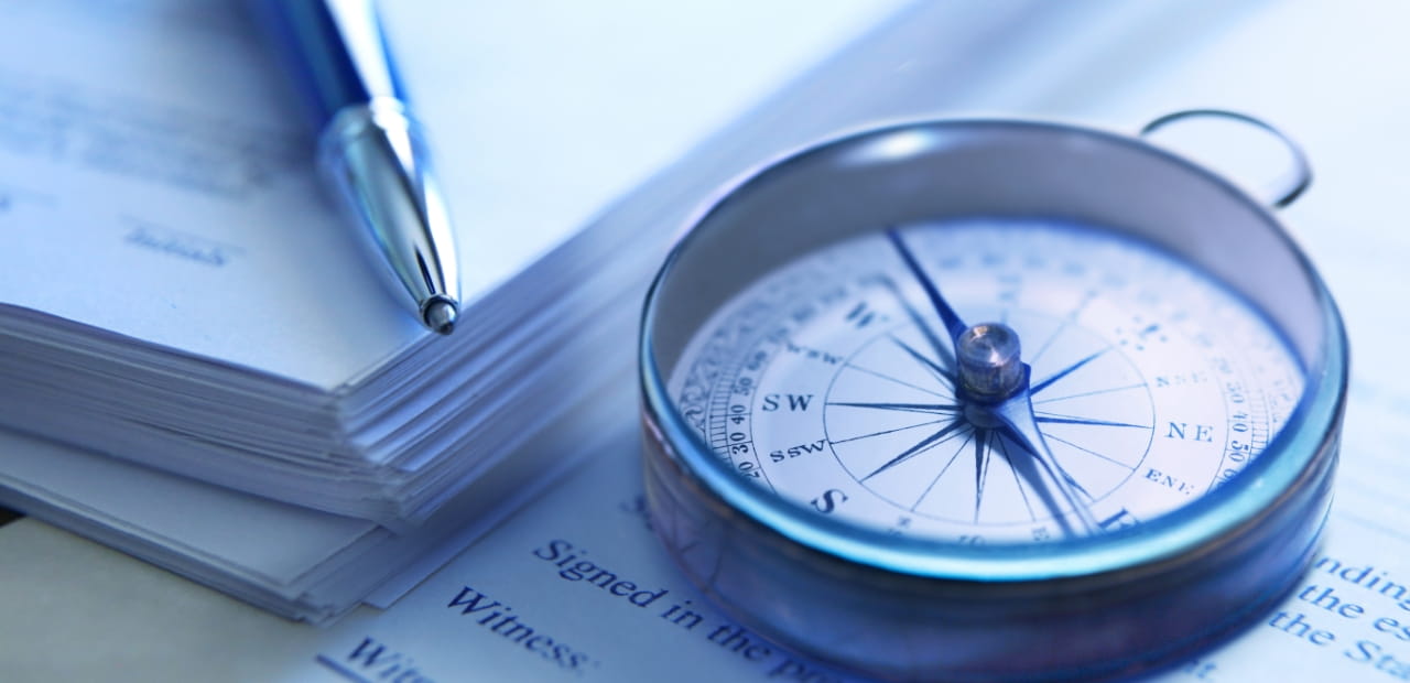 A close-up of a metal compass resting on documents beside a pen.