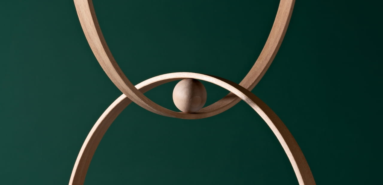 Two intersecting wooden rings with a small wooden sphere balanced at their meeting point, set against a dark green background.
